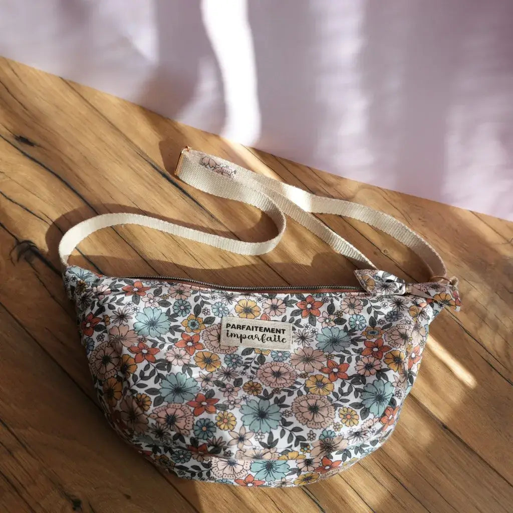 Sac banane demi-lune en tissu imprimé floral rétro, mêlant fleurs orange, bleues, jaunes et roses sur fond clair, posé sur une table en bois baignée de lumière naturelle. Doté d’une bandoulière réglable beige et d’une fermeture zippée, il présente une étiquette cousue “Parfaitement imparfaite” sur le devant. Accessoire pratique et élégant, idéal pour transporter l’essentiel avec une touche bohème et féminine.