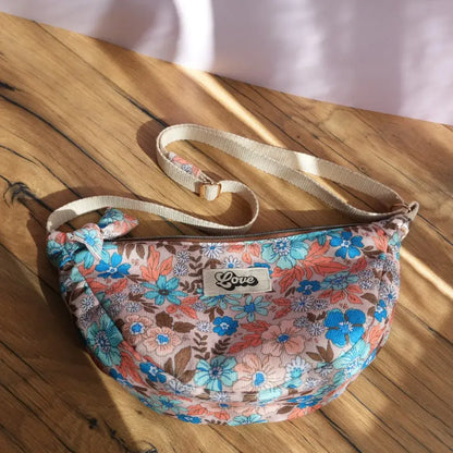 Sac banane demi-lune en tissu imprimé de fleurs hibiscus et feuillages, aux tons bleu turquoise, corail et marron, posé sur une table en bois éclairée par la lumière naturelle. Équipé d’une bandoulière réglable beige et d’une fermeture zippée, il arbore une étiquette centrale “Love” cousue sur le devant. Accessoire pratique et tendance, parfait pour transporter l’essentiel avec une touche estivale et romantique.