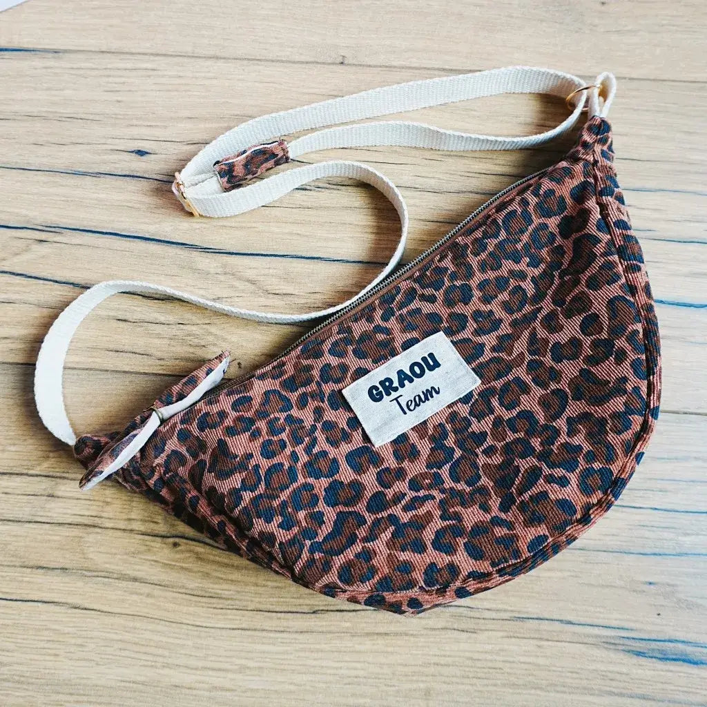 Sac banane demi-lune en tissu imprimé léopard dans des tons marron et noir, posé sur une table en bois clair. Doté d’une bandoulière réglable beige et d’une fermeture zippée, il présente une étiquette cousue sur le devant avec l’inscription “GRAOU Team”. Accessoire pratique et audacieux, idéal pour transporter ses affaires avec style et apporter une touche sauvage et tendance à une tenue.