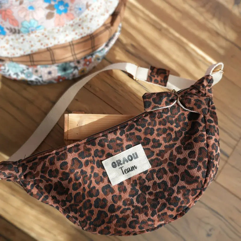 Sac banane demi-lune en tissu imprimé léopard marron et noir, avec bandoulière réglable beige et fermeture zippée, présenté sur une table en bois éclairée par la lumière naturelle. Une étiquette “GRAOU Team” est cousue sur le devant. À l’arrière-plan, on aperçoit partiellement d’autres sacs banane à motifs fleuris et à carreaux. Accessoire pratique et tendance, idéal pour un style audacieux et moderne.