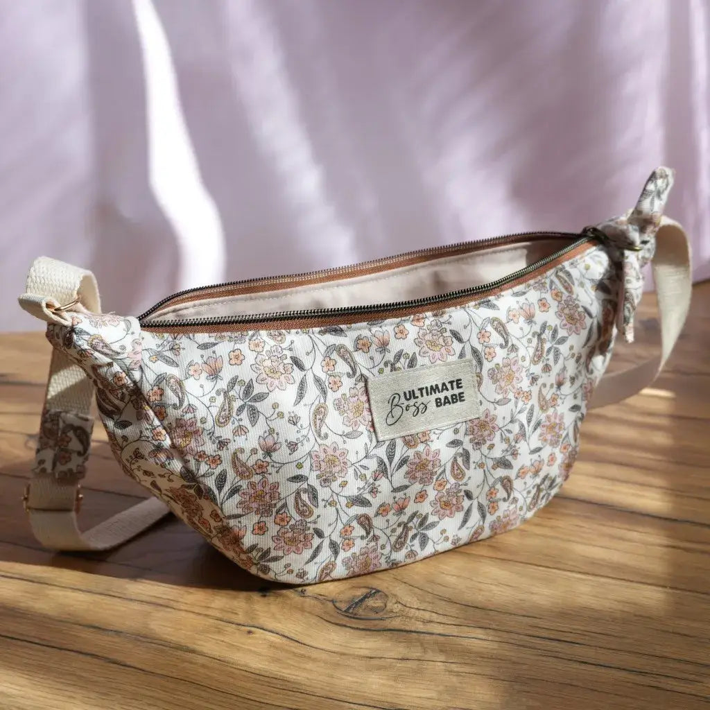 Sac banane demi-lune en velours milleraie clair décoré d’un motif floral délicat dans des teintes douces de rose, pêche et gris, disposé sur une table en bois baignée de lumière naturelle. Il possède une bandoulière ajustable beige, une fermeture zippée et une étiquette brodée “ULTIMATE Boss BABE” sur le devant. Un accessoire élégant et fonctionnel, parfait pour un look féminin et raffiné au quotidien.