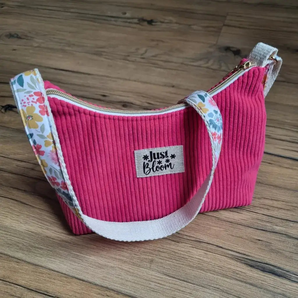 Sac bandoulière en velours côtelé framboise avec sangle en coton écru rehaussée d’un tissu fleuri coloré, doté d’une fermeture éclair dorée et d’une étiquette floquée 'Just Bloom'. Accessoire mode pratique et tendance, ce sac fait main combine élégance et originalité, idéal pour transporter ses affaires au quotidien tout en ajoutant une touche créative et personnalisée à son look.