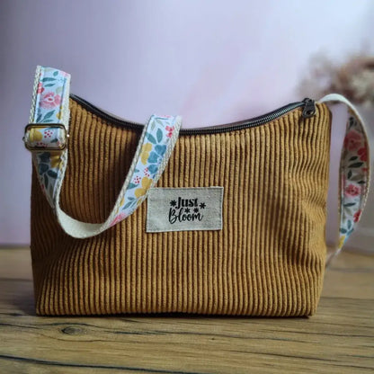 Sac bandoulière en velours côtelé jaune avec anse réglable en tissu fleuri coloré (bleu, rose, jaune), posé sur une surface en bois. Une étiquette "Just Bloom" est cousue sur le devant du sac. Fermeture zippée visible sur le dessus. Fond flou aux teintes roses et décor naturel en arrière-plan. Style bohème et fait main.