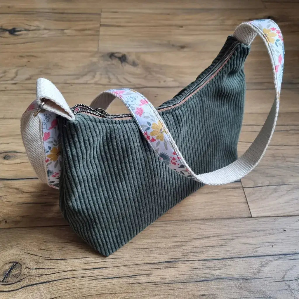 Sac bandoulière en velours côtelé vert avec fermeture zippée et sangle en coton ornée d’un motif fleuri coloré, posé sur un sol en bois. Ce sac au design moderne et naturel associe texture douce et imprimé floral pour un accessoire à la fois pratique et tendance, idéal au quotidien.