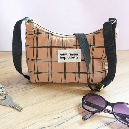 Sac bandoulière demi-lune en tissu à carreaux beige et noir, étiquette floquée 'Parfaitement imparfaite', anse réglable noire et zip métallique. Posé sur un parquet clair avec lunettes de soleil noires et trousseau de clés, cette mise en scène évoque un accessoire du quotidien pratique et stylé, parfait pour un look décontracté et tendance.