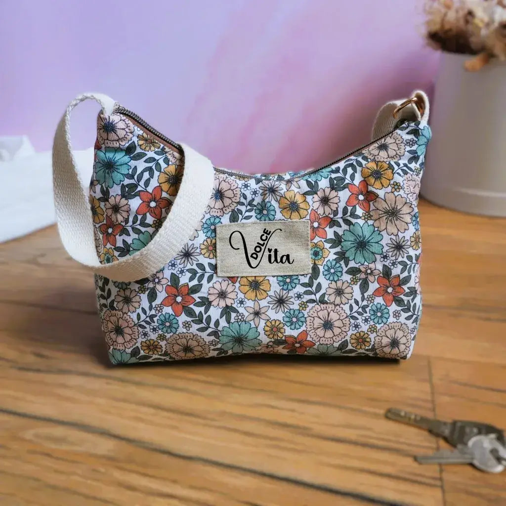 Sac bandoulière en tissu à motif fleuri rétro, mélange de fleurs rouges, orange, bleues et vertes sur fond clair, avec étiquette Dolce Vita et bandoulière écrue. Accessoire chic et romantique, idéal pour un look bohème et printanier. Présenté sur un sol en bois avec trousseau de clés et décor épuré pour un style cosy et tendance.