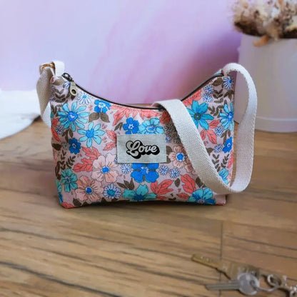 Sac bandoulière en tissu fleuri aux tons corail, turquoise et bleu, étiquette Love au centre, bandoulière écrue et zip doré. Ce sac féminin au style romantique et bohème est parfait pour accessoiriser vos tenues estivales. Mis en scène sur parquet clair avec clés et décor minimaliste, il incarne un look frais et tendance.
