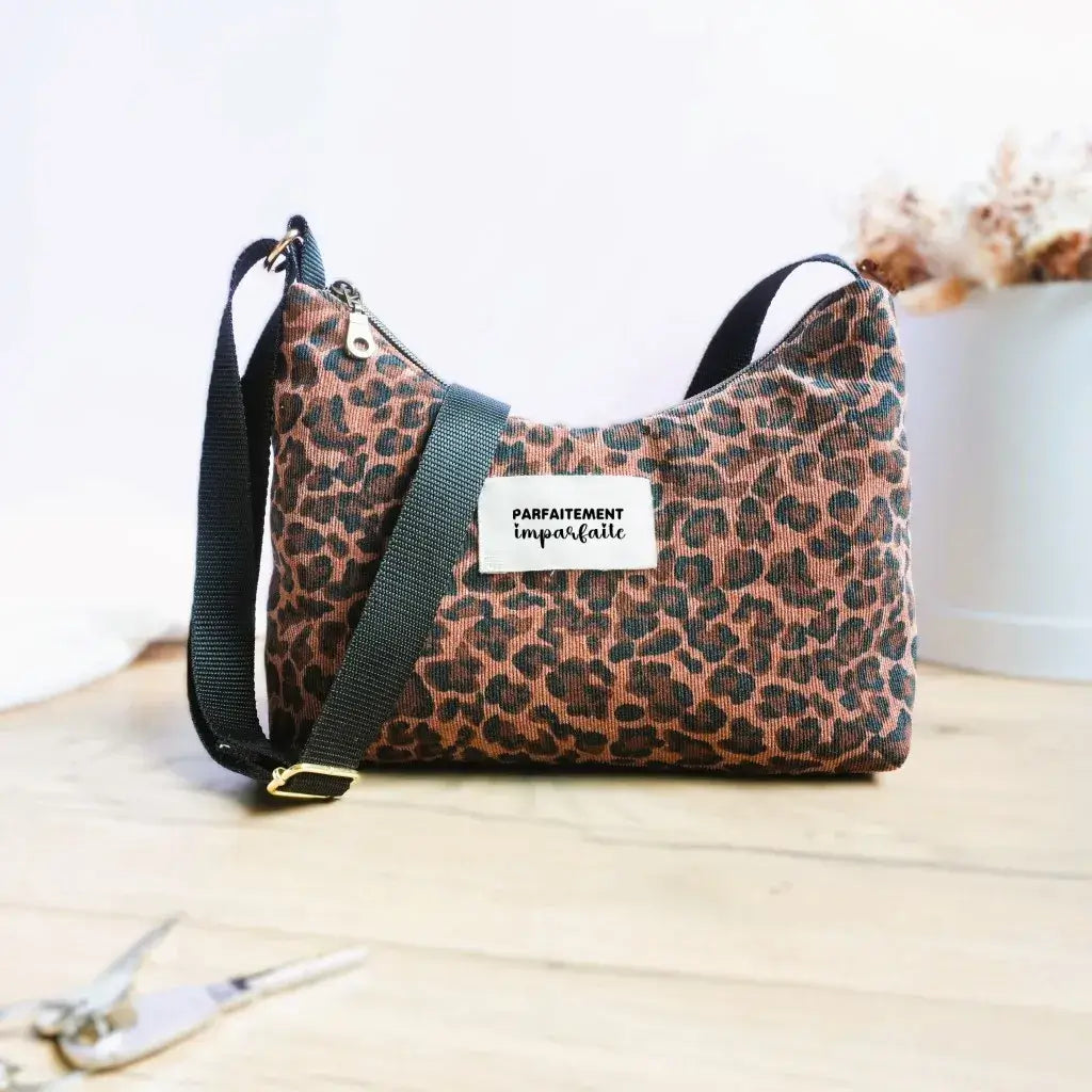 Sac bandoulière en velours à motif léopard brun et noir, anse réglable noire et étiquette 'Parfaitement imparfaite' floquée. Ce sac au style sauvage et tendance est idéal pour affirmer son look avec une touche audacieuse. Posé sur parquet clair avec trousseau de clés et décor minimaliste, parfait pour les sorties quotidiennes.