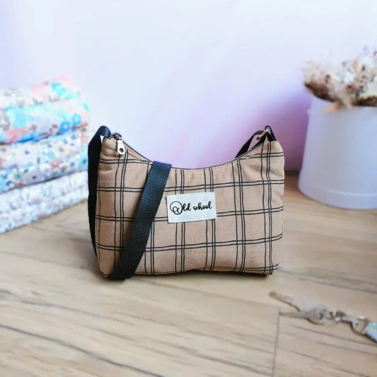 Sac bandoulière en tissu à carreaux beige et noir, style old school, avec anse réglable noire, posé sur un sol en bois clair. Accessoire tendance parfait pour un look vintage, idéal pour transporter téléphone, clés et petits objets au quotidien. Mise en scène déco avec pile de sacs identiques aux motifs fleuris  et vase de fleurs séchées en arrière-plan pour un rendu chaleureux.