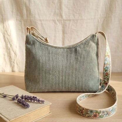 Sac bandoulière en velours côtelé vert lichen avec sangle fleurie, posé à côté d'un livre et de lavande.