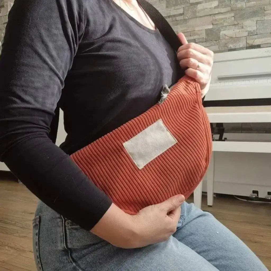 Sac demi-lune en velours côtelé de couleur terracotta. Ce sac est porté en bandoulière sur le côté par une demoiselle agenouillée et vêtue d'un t-shirt noir à manches longues et d'un jeans bleu. On distingue un piano blanc en arrière-plan.