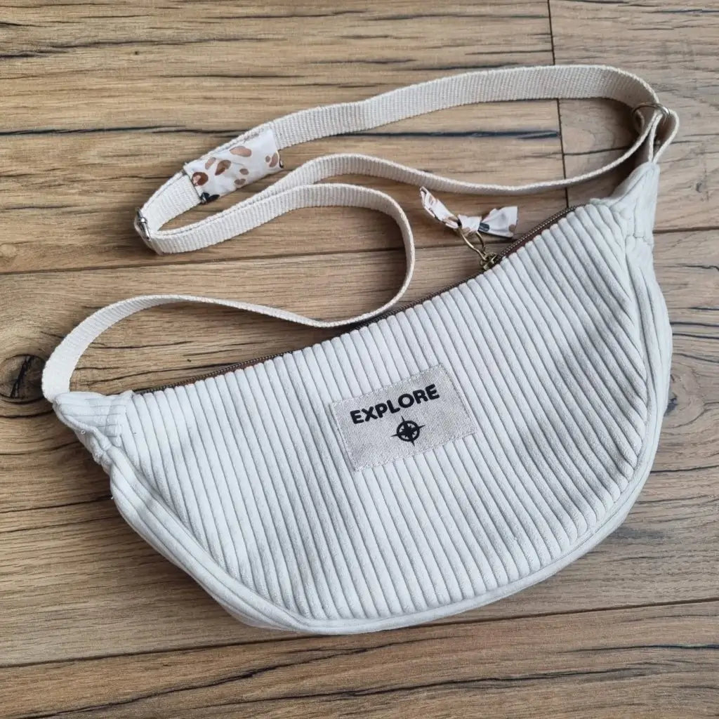 Sac demi-lune en velours côtelé de coloris beige avec une sangle beige posé au sol sur un parquet en chêne.