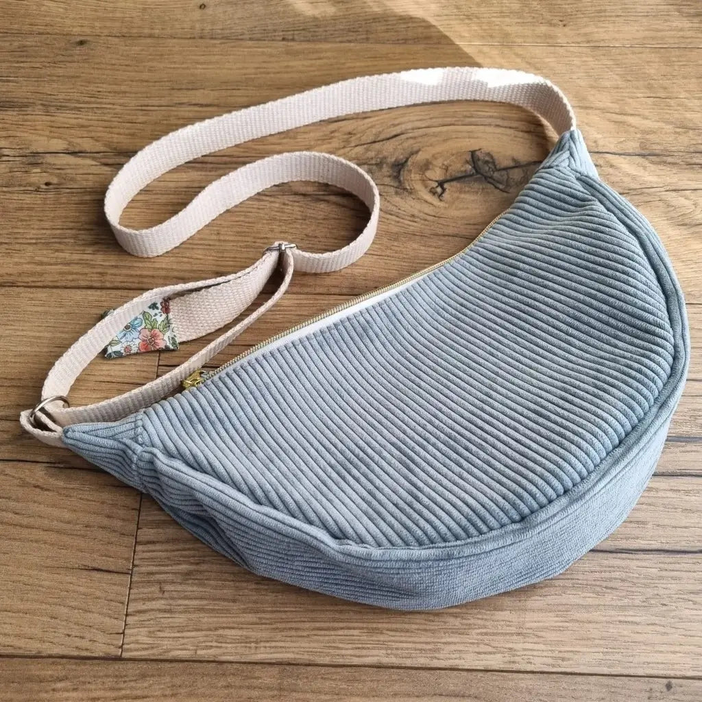 Sac demi-lune en velours côtelé de coloris bleu clair avec une sangle blanche posé au sol sur un parquet en chêne.