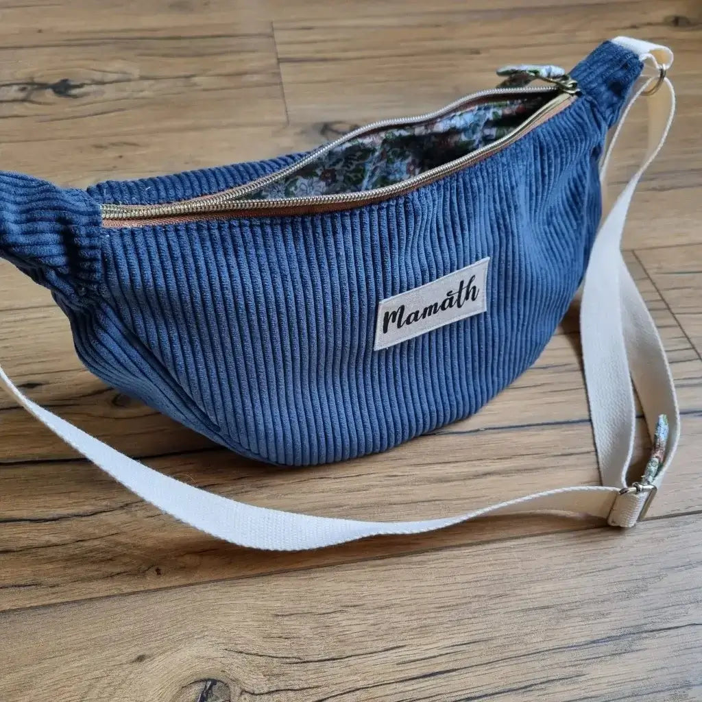 Sac demi-lune en velours côtelé de coloris bleu foncé avec une sangle blanche posé au sol sur un parquet en chêne. Ce sac est personnalisé avec une étiquette cousue à la main sur laquelle il est inscrit "Mamath".