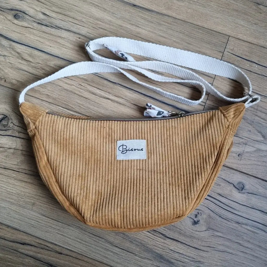 Sac demi-lune en velours côtelé de coloris jaune avec une sangle blanche posé au sol sur un parquet en chêne. Ce sac est personnalisé avec une étiquette cousue à la main sur laquelle il est inscrit "Bisous".