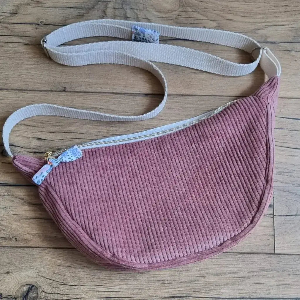 Sac demi-lune en velours côtelé de coloris rose avec une sangle blanche posé au sol sur un parquet en chêne.