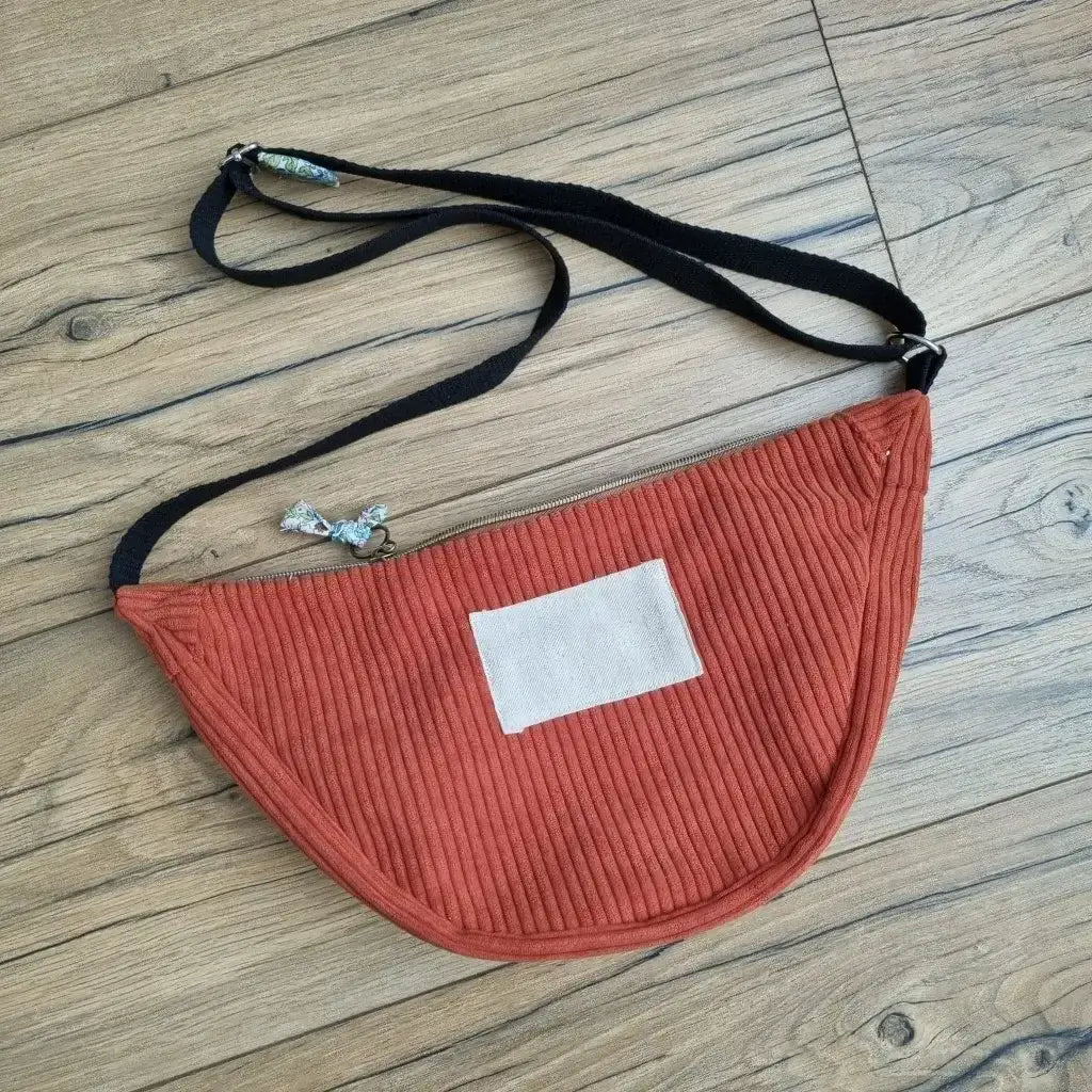 Sac demi-lune en velours côtelé de coloris terracotta avec une sangle noire posé au sol sur un parquet en chêne.