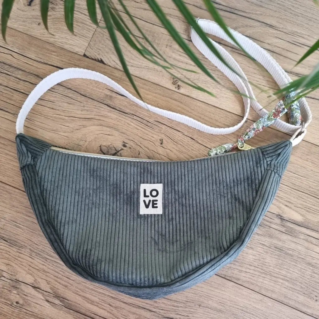 Sac demi-lune en velours côtelé de coloris vert avec une sangle blanche posé au sol sur un parquet en chêne. Ce sac est personnalisé avec une étiquette cousue à la main sur laquelle il est inscrit "LOVE".