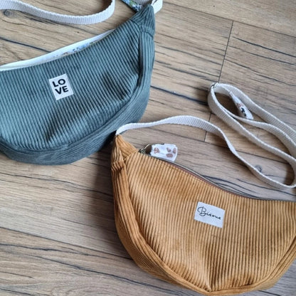 Deux sacs demi-lune à bandoulière posés au sol sur un parquet en chêne. En haut, le sac banane est en velours côtelé vert et personnalisé avec l'inscription "LOVE" floquée sur une étiquette en tissu. Celui du bas est de couleur jaune avec une personnalisation "Bisous" en façade.