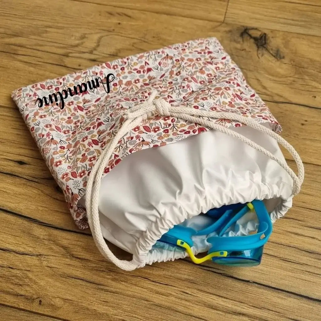 Sac piscine imperméable au format pochon se fermant grâce à deux cordelettes en coton blanc de part et d'autre et cousu avec un tissu imprimé à motif liberty. Le sac est personnalisé avec un prénom floqué.