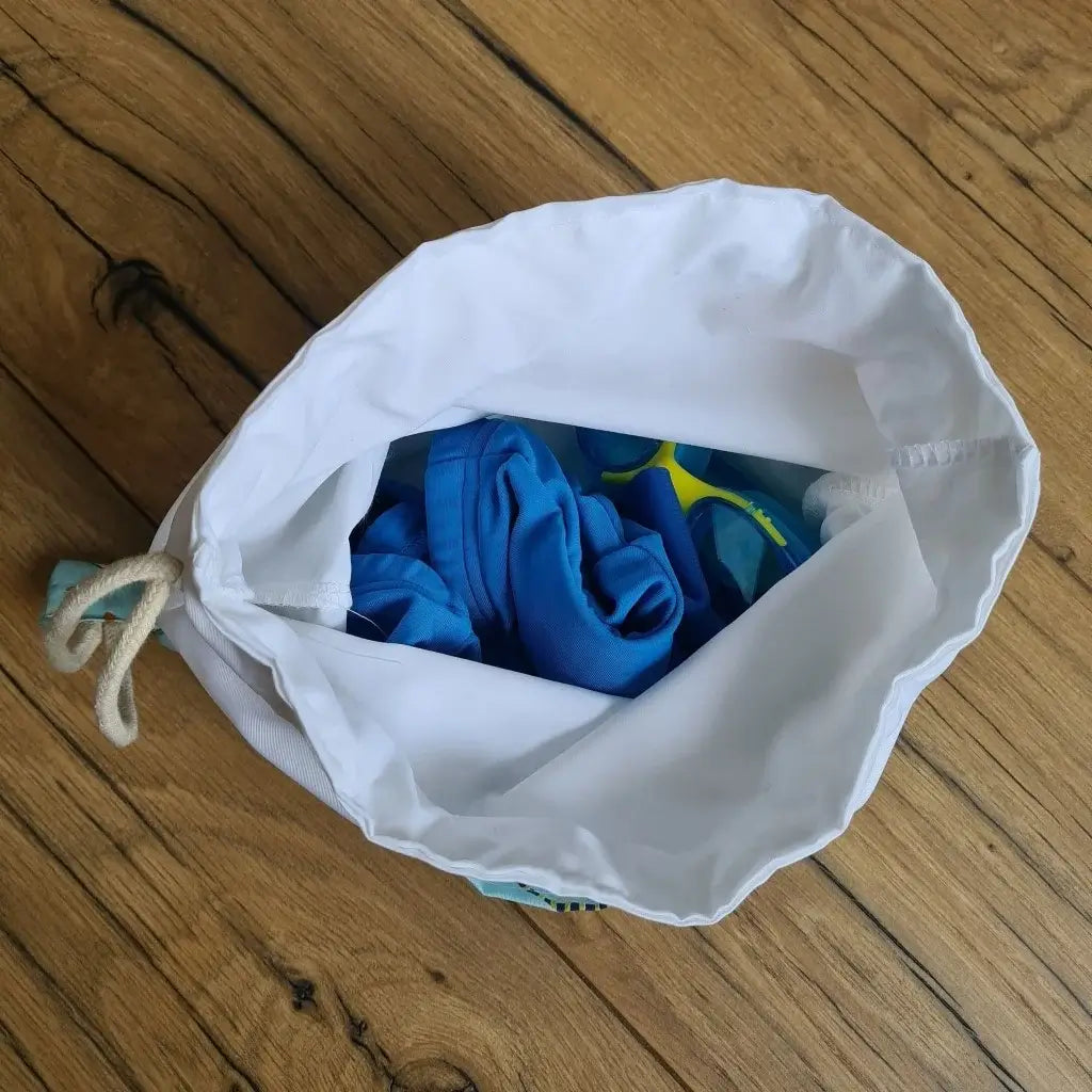 Vue du dessus d'un sac piscine imperméable au format pochon montrant la doublure intérieur en tissu imperméable et l'intérieur du sac comprenant un maillot de bain bleu et des lunettes de piscine.