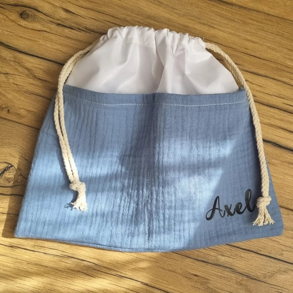 Sac piscine imperméable au format pochon se fermant grâce à deux cordelettes en coton blanc de part et d'autre et cousu avec un tissu en double gaze de coton uni de coloris bleu jeans. Le sac pochon est personnalisé avec le prénom "Axel" floqué en bas à droite.