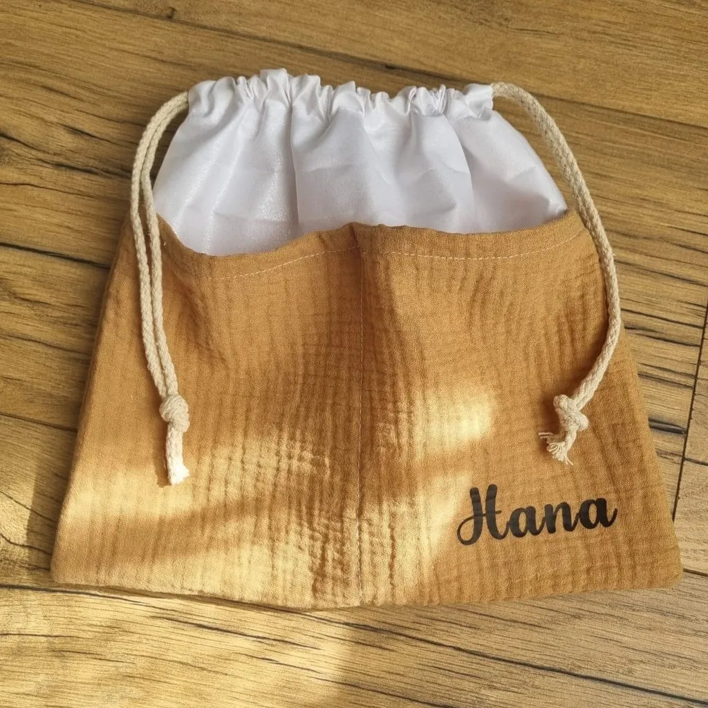 Sac piscine imperméable au format pochon se fermant grâce à deux cordelettes en coton blanc de part et d'autre et cousu avec un tissu en double gaze de coton uni de coloris camel. Le sac pochon est personnalisé avec le prénom "Hana" floqué en bas à droite.