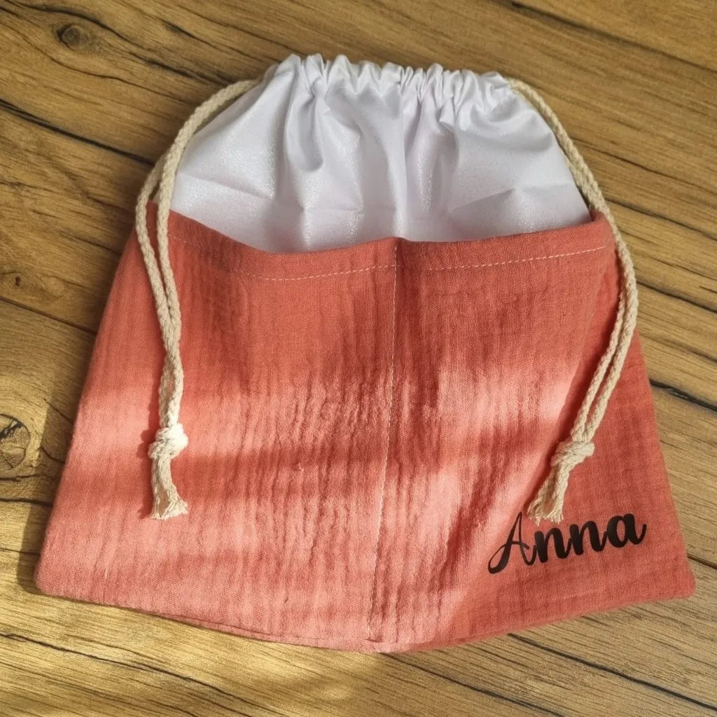 Sac piscine imperméable au format pochon se fermant grâce à deux cordelettes en coton blanc de part et d'autre et cousu avec un tissu en double gaze de coton uni de coloris marsala. Le sac pochon est personnalisé avec le prénom "Anna" floqué en bas à droite.