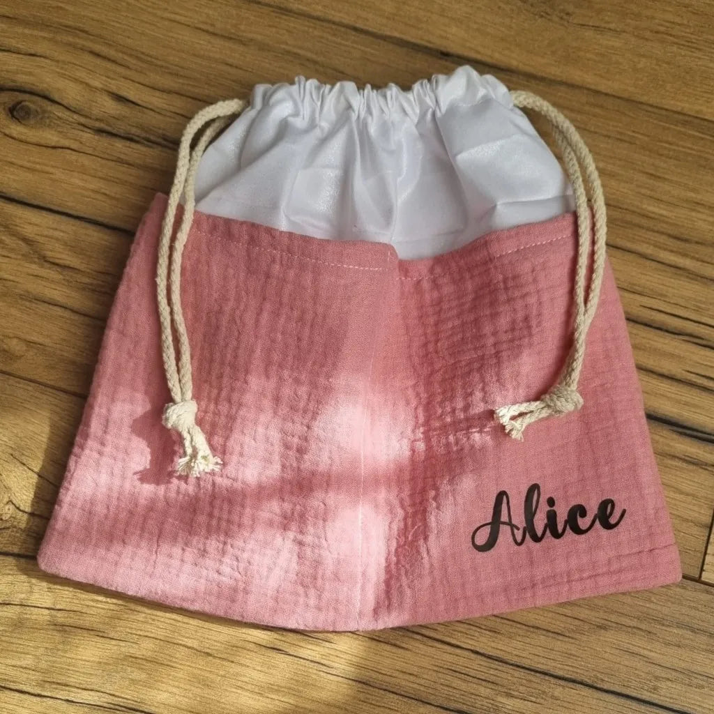 Sac piscine imperméable au format pochon se fermant grâce à deux cordelettes en coton blanc de part et d'autre et cousu avec un tissu en double gaze de coton uni de coloris rose. Le sac pochon est personnalisé avec le prénom "Alice" floqué en bas à droite.