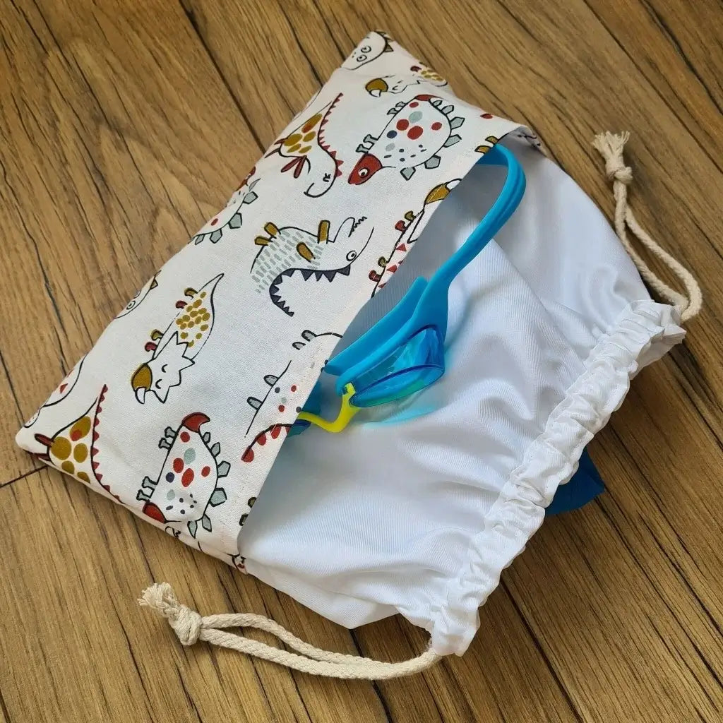 Sac piscine imperméable au format pochon se fermant grâce à deux cordelettes en coton blanc de part et d'autre et cousu avec un tissu imprimé à motif dinosaures.