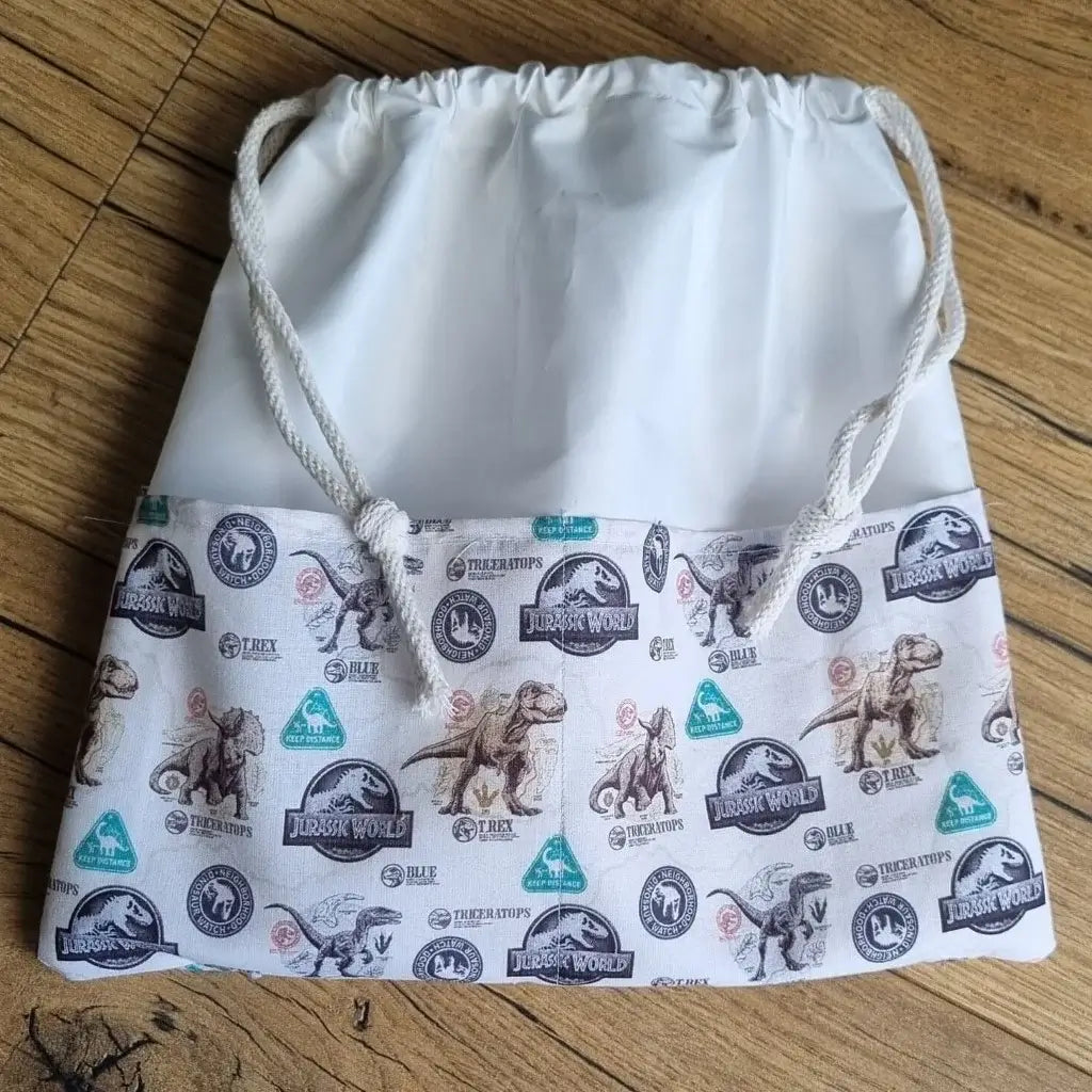 Sac piscine imperméable au format pochon se fermant grâce à deux cordelettes en coton blanc de part et d'autre et cousu avec un tissu imprimé à motif de dinosaures de jurassic park.