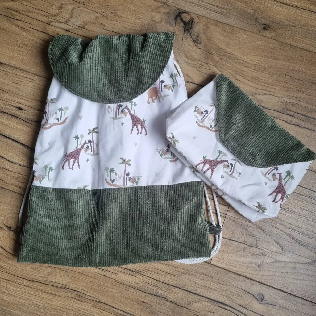 Sac pochon pour enfant avec une base et un rabat en velours côtelé vert. Le corps du sac est en tissu avec un motif savane. A droite du sac se trouve une petite pochette assortie, également avec un tissu motif savane et un rabat en velours côtelé vert.