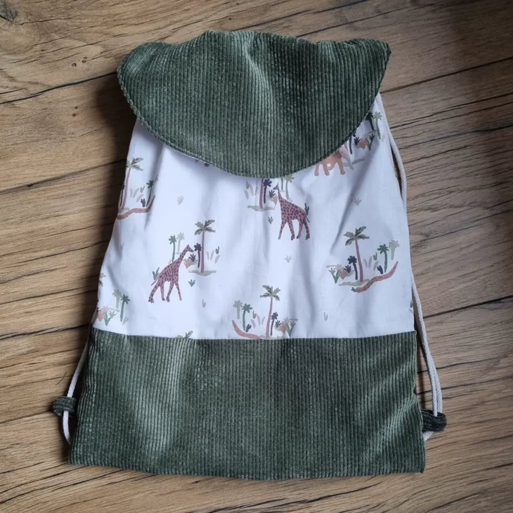 Sac pochon pour enfant posé au sol sur un parquet en chêne. La base du sac et son rabat sont en velours côtelé vert foncé. Le corps du sac est en tissu avec un motif savane.