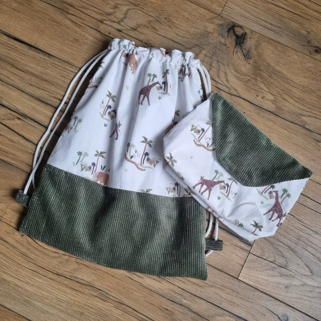 Sac pochon pour enfant avec une base en velours côtelé vert. Le corps du sac est en tissu avec un motif savane. A droite du sac se trouve une petite pochette assortie, également avec un tissu motif savane et un rabat en velours côtelé vert.