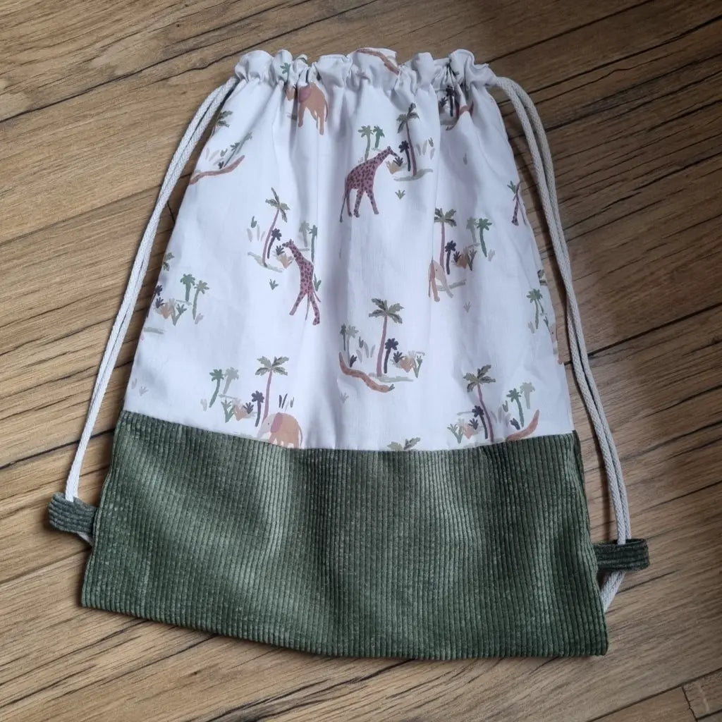 Sac pochon pour enfant posé au sol sur un parquet en chêne. La base du sac est en velours côtelé vert foncé. Le corps du sac est en tissu avec un motif savane.