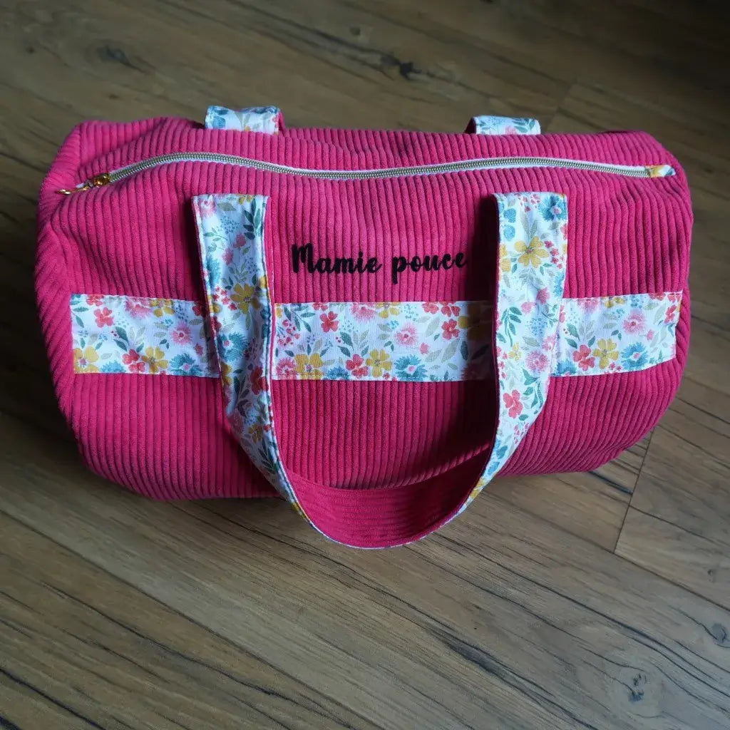 Sac polochon en velours côtelé coloris framboise, orné de larges anses et de bandes décoratives en coton imprimé floral multicolore. Personnalisé avec l’inscription “Mamie pouce” floquée en velours noir sur le devant, il est équipé d’une fermeture éclair dorée sur le dessus. Posé sur un sol en bois, ce sac artisanal allie praticité et style, idéal comme bagage de voyage ou sac de sport personnalisé.