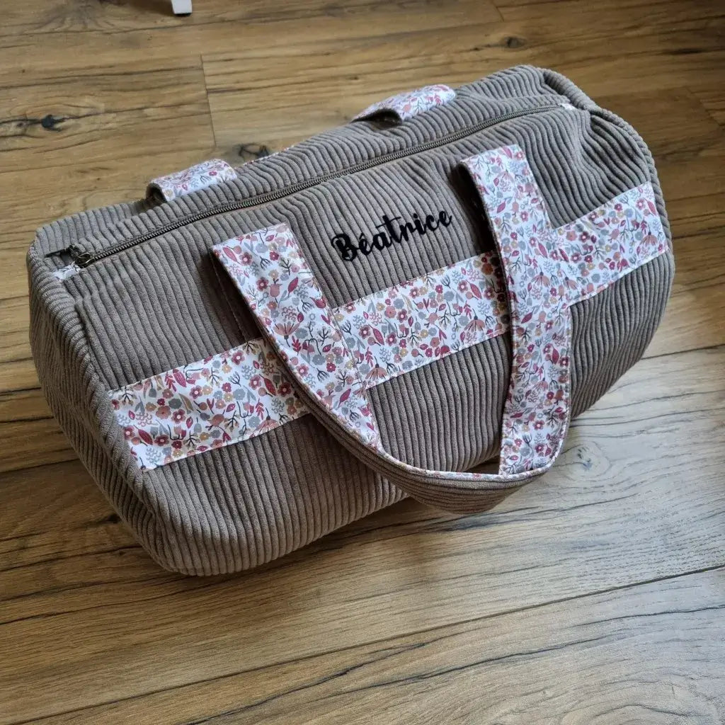 Sac polochon personnalisé avec le prénom "Béatrice", confectionné en velours côtelé taupe, posé sur un sol en bois. Les anses et la bande décorative sont réalisées dans un tissu fleuri rose, rouge et blanc, apportant une touche délicate et féminine. Fermeture zippée sur le dessus. Idéal comme sac de voyage, sac week-end ou sac de maternité fait main au style élégant et pratique.