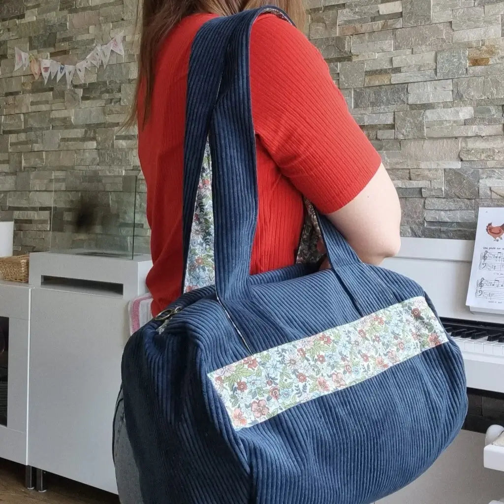 Sac polochon en velours côtelé bleu foncé avec une bande en tissu coton imprimé fleuri porté en bandoulière par une femme vêtue d'un t-shirt rouge.