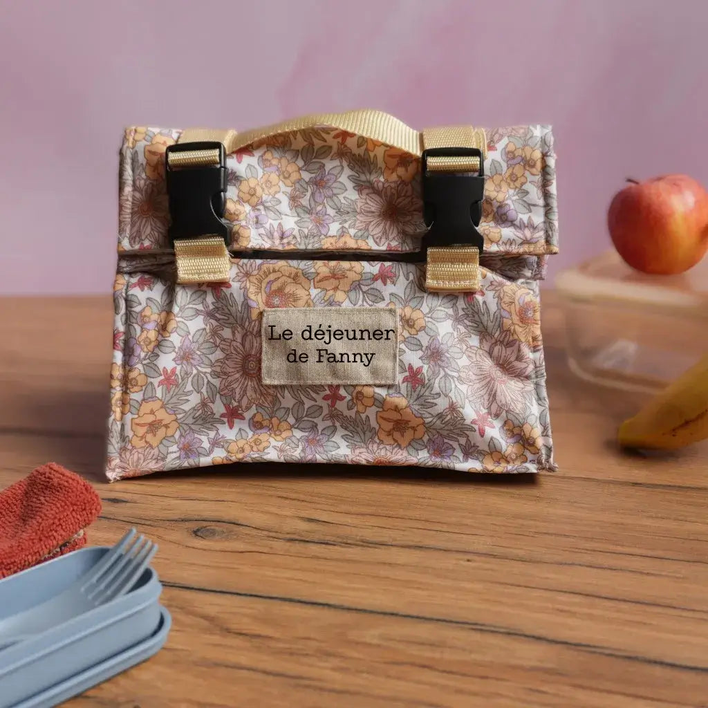 Sac repas isotherme en coton enduit fleuri, avec inscription personnalisée “Le déjeuner de Fanny”, équipé d’une poignée solide et de fermetures à clips noires et sangles beiges. Lunch bag artisanal élégant et pratique, idéal pour transporter le repas au travail, à l’école ou en pique-nique, tout en conservant les aliments à bonne température. Accessoire réutilisable, durable et au design tendance.