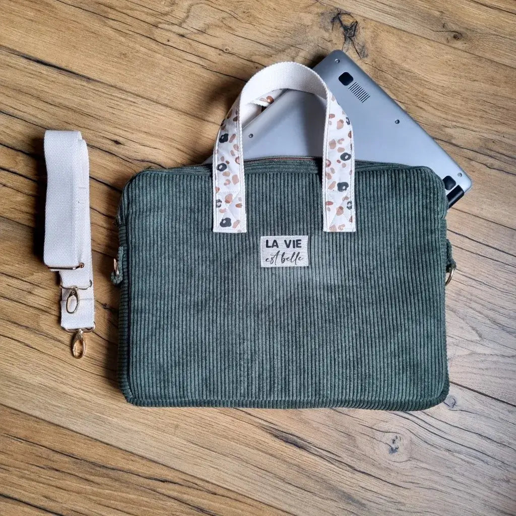 Sacoche à bandoulière pour ordinateur portable en velours côtelé de coloris vert avec une doublure en coton imprimé au motif léopard. Cette sacoche est personnalisable avec une inscription de son choix floquée sur une étiquette en tissu (ici, "La vie est belle"). Une bandoulière blanche est enroulée à gauche de la sacoche. Un ordinateur portable est glissé à l'intérieur de celle-ci.