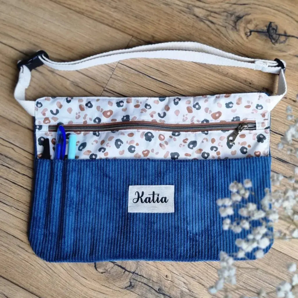 Tablier ceinture en velours côtelé bleu foncé, personnalisable avec un prénom floqué sur une étiquette en tissu, doté d'un coton imprimé léopard et de multiples compartiments pratiques pour stylos et petits accessoires. Idéal pour les créatrices, couturières ou pour offrir à des maîtresses qui veulent allier praticité et élégance au quotidien.