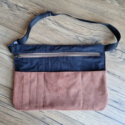 Tablier-ceinture pratique en velours côtelé marron associé à une partie supérieure en tissu noir uni, équipé d’une grande poche zippée sur le devant. Il dispose d’une sangle réglable noire pour être porté à la taille ou en bandoulière. Posé sur un sol en bois, cet accessoire artisanal allie fonctionnalité et style, idéal pour les créateurs, artisans ou vendeurs souhaitant garder leurs outils ou accessoires à portée de main.