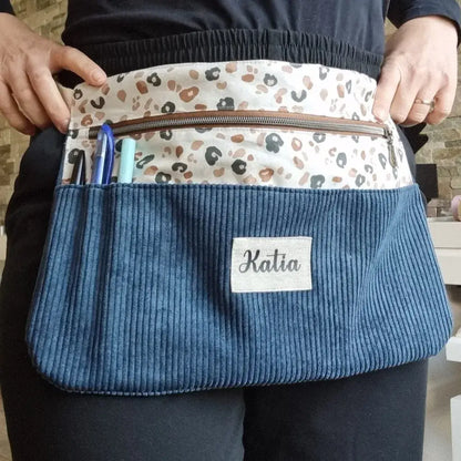 Tablier ceinture en velours côtelé bleu porté à la taille, personnalisé avec un prénom floqué 'Katia', et doté d’un imprimé léopard coloré. Ce modèle pratique comporte plusieurs poches de rangement pour stylos ou outils, idéal pour les créatrices, couturières ou professionnelles du fait main.