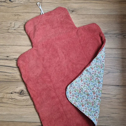 Tapis à langer nomade et imperméable en éponge de bambou de coloris terracotta. Ce tapis est replié sur l'un de ses côté pour laisser apparaître l'envers composé d'un tissu en coton imprimé à motif fleuri. Ce tapis dispose également d'un cordon élastique blanc à l'une de ses extrémités pour pouvoir le maintenir une fois enroulé.