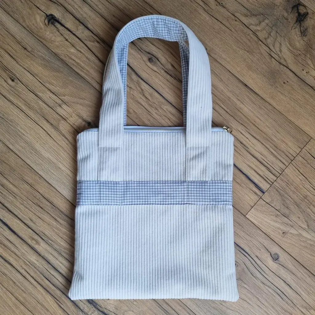 Sac fourre-tout de type tote-bag en velours côtelé de coloris beige. Le sac présente en façade une bande horizontale en tissu à motif carreaux beiges. Ce même tissu se retrouve également au dos des deux anses.