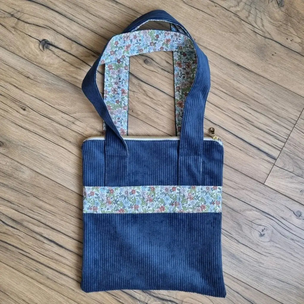 Sac fourre-tout de type tote-bag en velours côtelé de coloris bleu foncé. Le sac présente en façade une bande horizontale en tissu à motif fleuri. Ce même tissu se retrouve également au dos des deux anses.