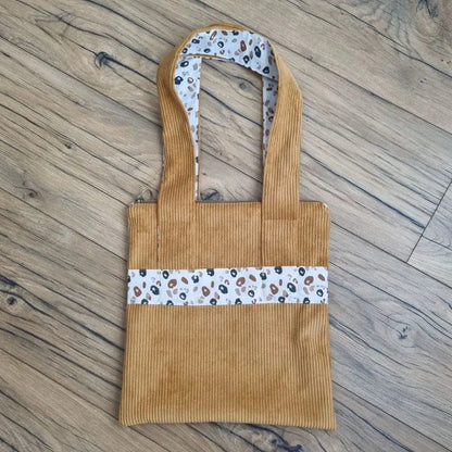 Sac fourre-tout de type tote-bag en velours côtelé de coloris jaune. Le sac présente en façade une bande horizontale en tissu à motif léopard. Ce même tissu se retrouve également au dos des deux anses.