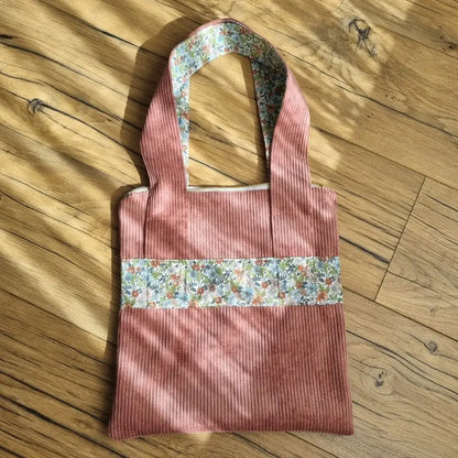Sac fourre-tout de type tote-bag en velours côtelé de coloris rose. Le sac présente en façade une bande horizontale en tissu à motif fleuri. Ce même tissu se retrouve également au dos des deux anses.