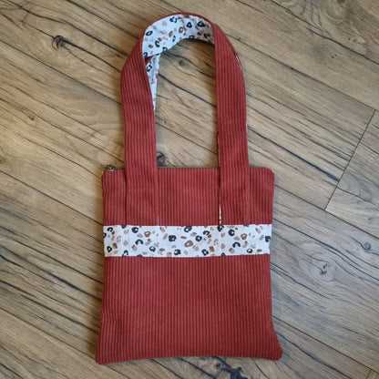Sac fourre-tout de type tote-bag en velours côtelé de coloris terracotta. Le sac présente en façade une bande horizontale en tissu à motif léopard. Ce même tissu se retrouve également au dos des deux anses.