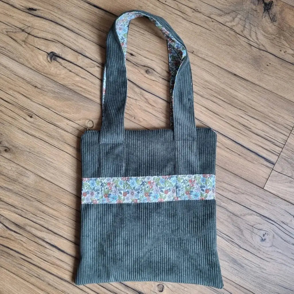 Sac fourre-tout de type tote-bag en velours côtelé de coloris vert. Le sac présente en façade une bande horizontale en tissu à motif fleuri. Ce même tissu se retrouve également au dos des deux anses.